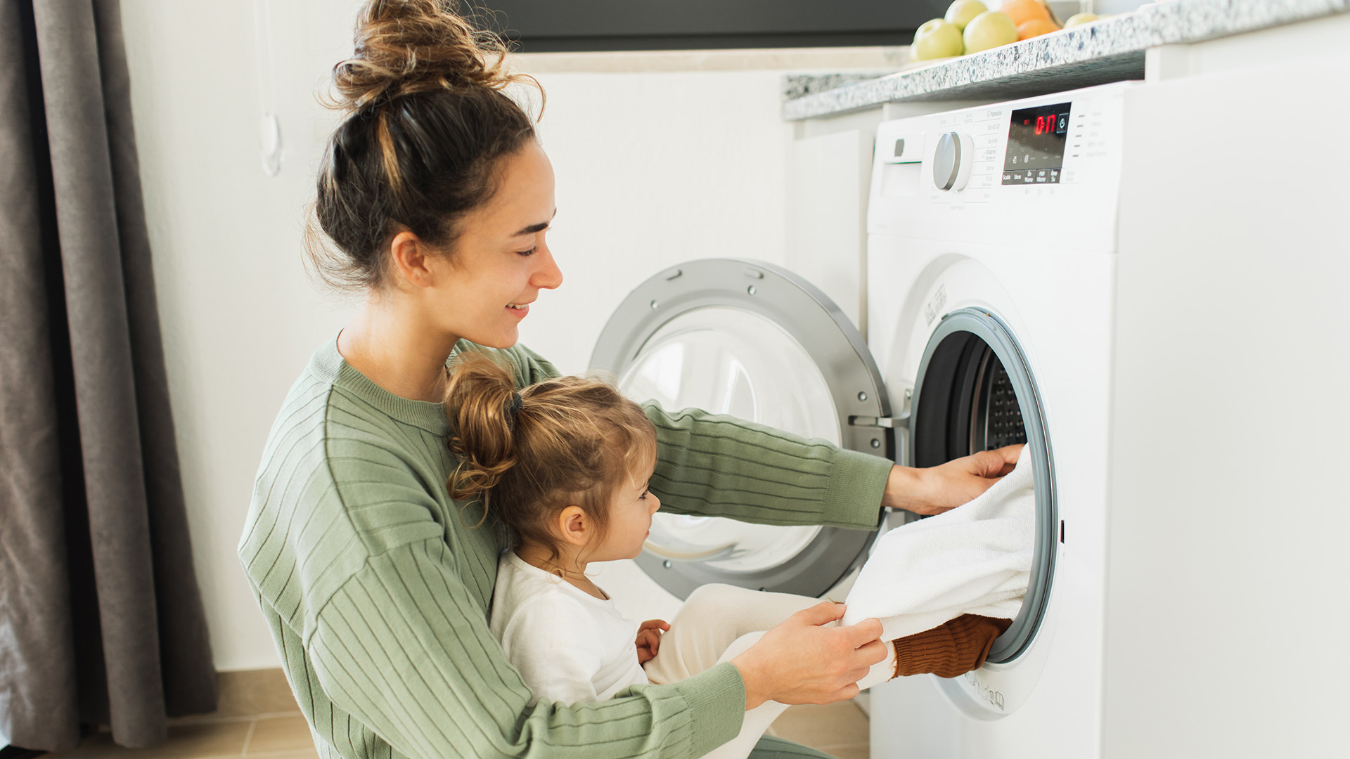 Mother a housewife with a baby engaged in laundry fold clothes into the washing machine. Beautiful mother and her daughter enjoying and laughing doing the laundry in the kitchen of modern apartment where a washing machine can be seen on.